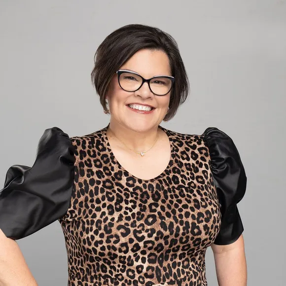 Woman with dark hair and glasses smiles, wearing leopard-print top with black puffy sleeves.
