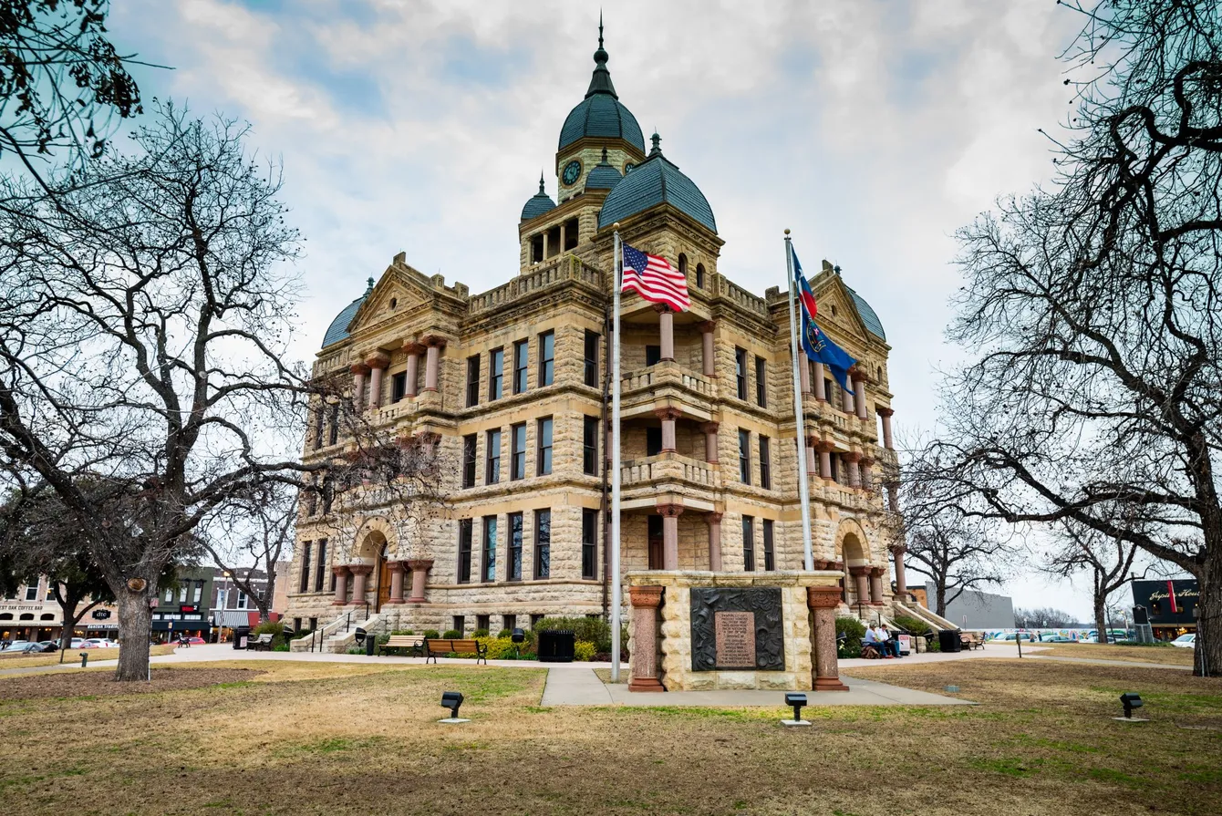 Historic Denton County Courthouse, Texas, beige stone building, dome, flags, trees, cloudy sky.