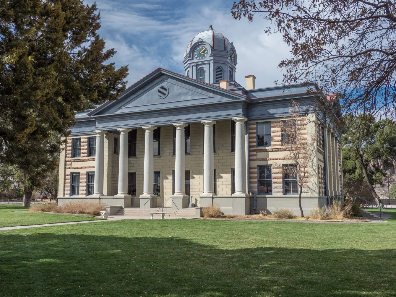 Historic courthouse with columns and a dome, on a green lawn with trees under a partly cloudy sky.
