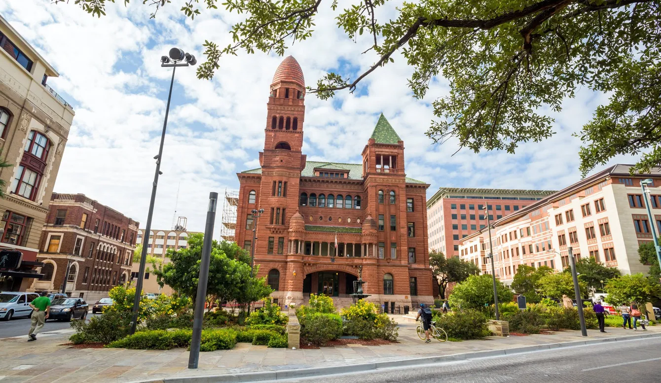 Red brick building with tower and green roof, in an urban setting with trees and a cloudy sky.
